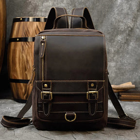 The Leather Travel Daypack Shoulder Bag, made from genuine cowhide leather in dark brown, is showcased against a rustic backdrop featuring a wooden barrel. This versatile daypack includes brass hardware, two buckled front straps, a vertical zipper pocket, and padded shoulder straps. Its overall style exudes vintage ruggedness.