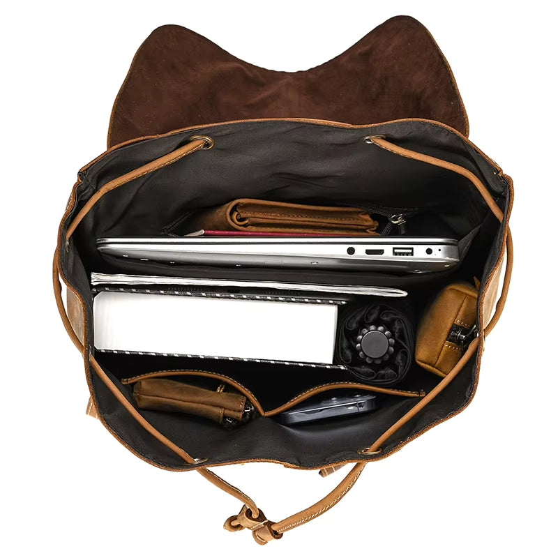 A top view of the Vintage Leather Backpack reveals travel essentials neatly arranged inside: a laptop, notebook, wallet, smartphone, and a small pouch. Made from genuine cowhide leather, this backpack features adjustable straps and is displayed open to showcase its contents.