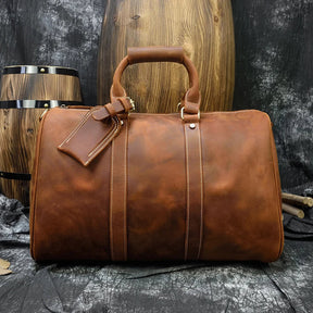 A large brown Weekender 18 Leather Bag, made from genuine cowhide leather, is positioned against a rustic wooden backdrop with wooden barrels. The bag features a top handle, spacious interior pockets, a secured tag, and visible stitching that adds to its vintage appeal.