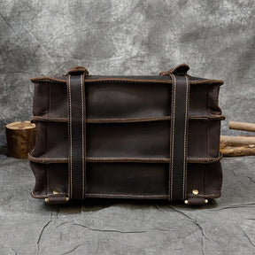 A large, dark brown Leather Business Travel Briefcase Backpack with visible stitching is set against a textured gray background. The backpack features multiple horizontal straps and brass rivets at the corners, showcasing a rugged and durable design. Logs are partially visible in the background.