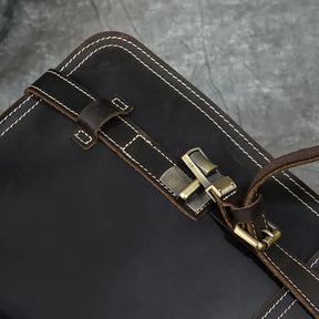 Close-up of the Leather Business Travel Briefcase Backpack crafted from genuine dark brown cowhide leather with white stitching. This versatile bag features a brass buckle and clasp on its strap, set against a textured grey background.