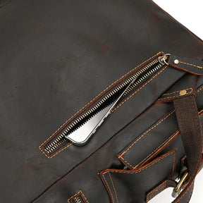 A close-up of the Genuine Leather Travel Backpack showcases a dark brown leather exterior with visible orange stitching. The backpack features an open zipper pocket where a shiny silver comb can be partially seen inside. A portion of a dark brown strap is also visible on the right side.