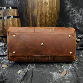 A Weekender 18 Leather Bag, crafted from genuine cowhide and adorned with metal studs on the bottom, rests on a textured gray surface. Wooden barrels and a large wooden panel provide a rustic backdrop, with fallen leaves scattered around to complete the scene. The bag boasts spacious interior pockets for added convenience.