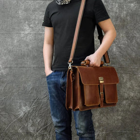 A person dressed in a black t-shirt, blue jeans, and a patterned scarf carries the Leather 16" Business Briefcase with an adjustable strap made of genuine cowhide leather over their shoulder and holds its handles with one hand. The background is a textured gray surface.