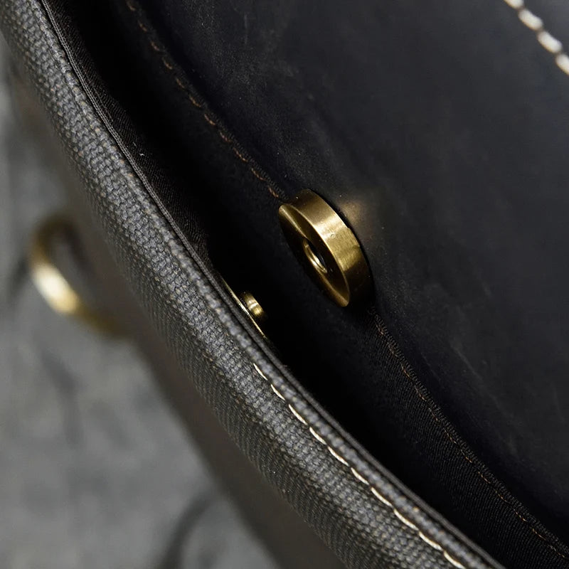 Close-up view of a dark-colored Leather Business Travel Briefcase Backpack with a brass-colored magnetic button clasp. The image focuses on the interior lining and the stitching along the edge of the bag flap. This genuine cowhide leather briefcase showcases meticulous craftsmanship.