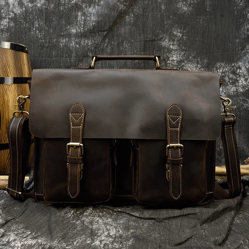 The Leather 16" Business Briefcase, a stylish accessory crafted from genuine cowhide leather in dark brown, is showcased against a gray, textured backdrop. This elegant satchel includes brass buckles and fittings, a top handle for easy carrying, two front pockets with secure strap closures, and an adjustable shoulder strap for versatility. A wooden barrel can be seen in the background.