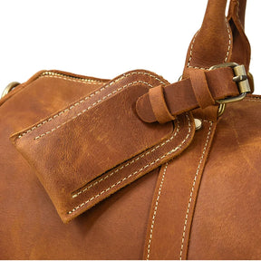 Close-up of a brown leather bag with visible stitching. The image highlights a buckle and a strap, showcasing the durable Genuine Cowhide Leather texture and craftsmanship details. This Weekender 24 Duffle Bag has a rustic, well-worn appearance perfect for travellers seeking style and durability.