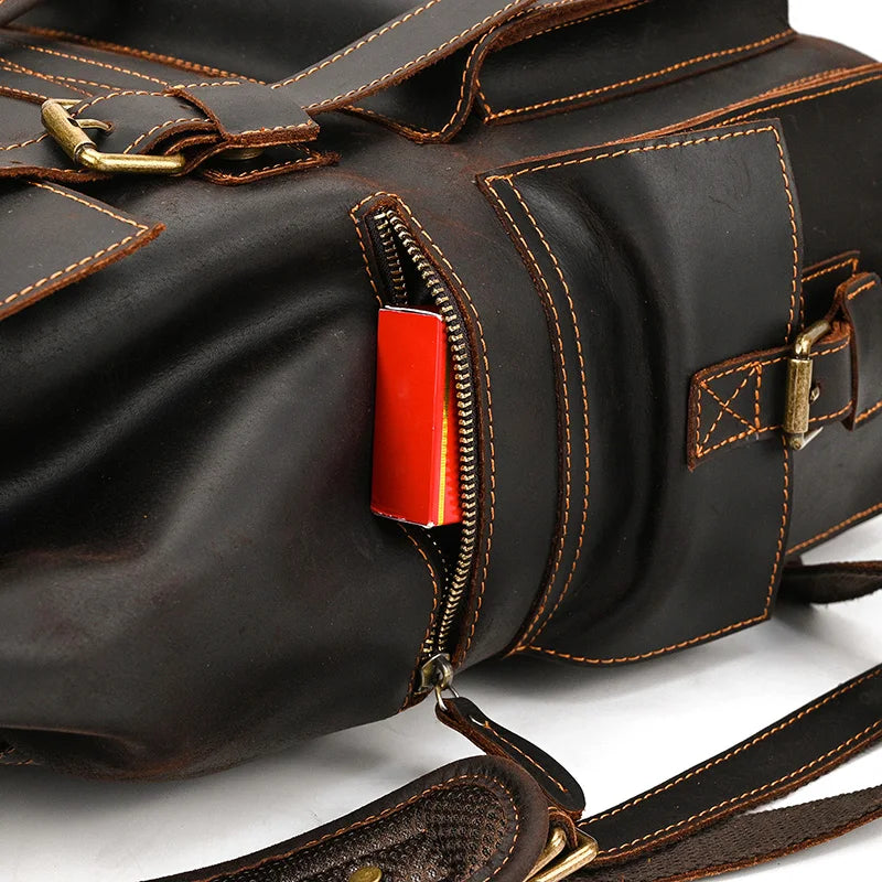 A close-up shot of the Genuine Leather Travel Backpack in dark brown showcases its brass hardware and distinctive white stitching. The side pocket is unzipped, revealing a red rectangular object inside. The leather exhibits a high-quality, rugged, and stylish texture.