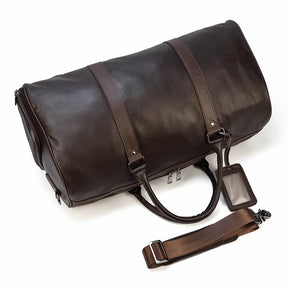 The Weekender Duffle 21 Leather Travel Bag is a dark brown genuine leather travel bag featuring dual handle straps and a detachable shoulder strap. It includes metal clasps, a zippered section, and a clear ID tag holder on the front. The bag is positioned at a slight angle on a white background.