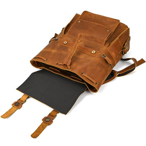 The Genuine Leather Travel Backpack is showcased with its front flap open, unveiling a black interior. This vintage-style backpack boasts multiple pockets, brass hardware, and adjustable straps. The front flap is secured with two leather straps featuring buckles.