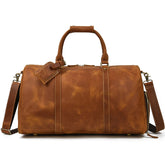 A stylish brown Weekender 24 Duffle Bag crafted from genuine cowhide leather, featuring a handle on top and a detachable shoulder strap. Perfect for travelers, the bag includes a luggage tag attached to one of the handles and brass hardware detailing.