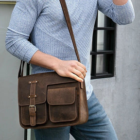 A person wearing a light blue long-sleeve shirt and denim jeans is carrying a Leather Business Travel Shoulder Bag, which is brown with multiple pockets and alloy hardware. The person is standing in front of a window with black trim.