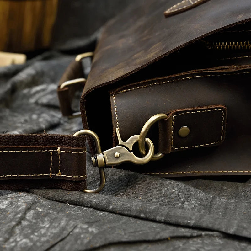 Close-up of the Leather 16" Business Briefcase showcasing its brass hardware and stitching details. The focus is on the side of the briefcase, where the adjustable strap connects to the bag via a metal hook and loop. The genuine cowhide leather briefcase rests on a textured, dark fabric background.