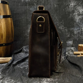 A side view of the Leather 16" Business Briefcase, crafted from dark brown genuine cowhide leather, showcasing a metal ring attachment, stitching details, and a brass buckle. The briefcase features an adjustable strap and is set against a textured dark gray backdrop with a wooden barrel and chopped logs partially visible in the background.