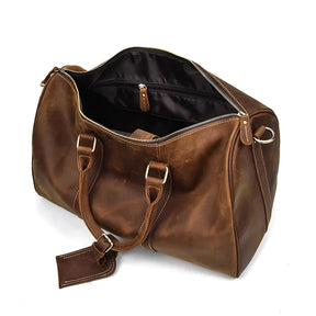 The Weekender 18 Leather Bag is a spacious brown duffel crafted from genuine cowhide leather. It features a roomy interior with an open main compartment and black inner lining. The bag comes with sturdy handles, metal hardware, and an attached luggage tag. A partially open zipper reveals ample space along with interior pockets for additional storage.