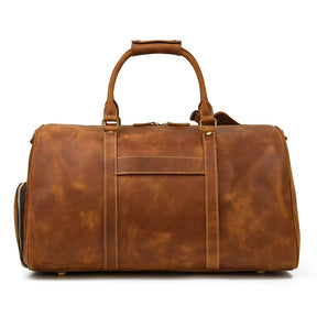 A large, stylish Weekender 24 Duffle Bag made of genuine cowhide leather is shown. With sturdy handles and reinforced stitching, this rectangular bag features antique brass hardware, a front zip pocket, and padded straps for carrying—ideal for travelers.