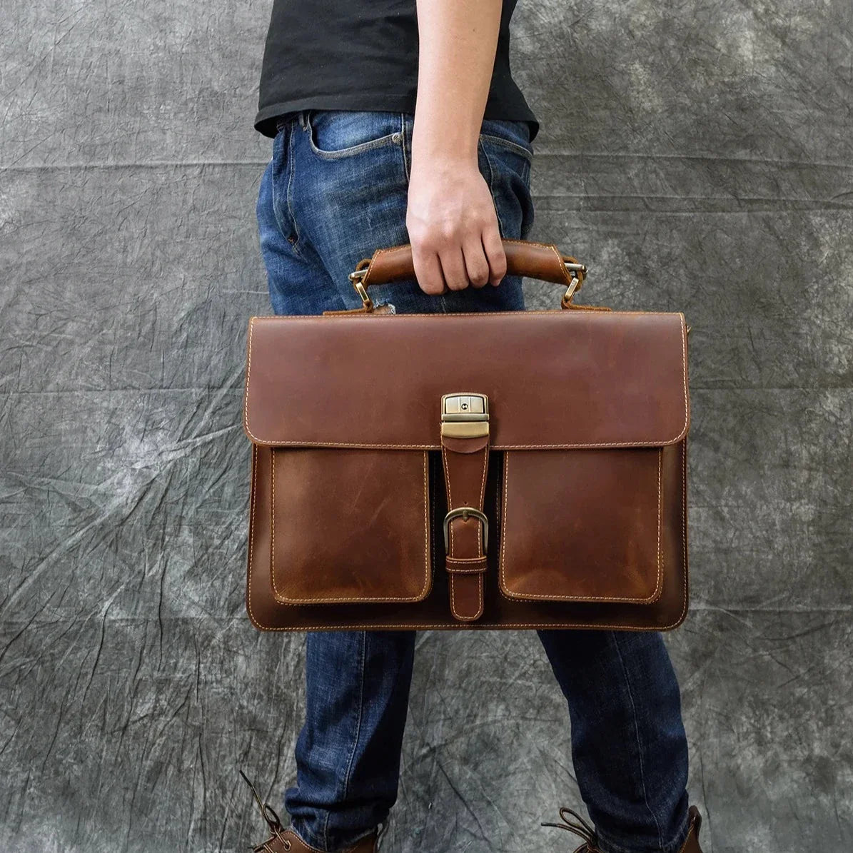 A person wearing jeans and a black shirt holds the Leather 16" Business Briefcase, crafted from genuine cowhide leather and featuring two front pockets with a buckle clasp. The background is a textured gray backdrop.