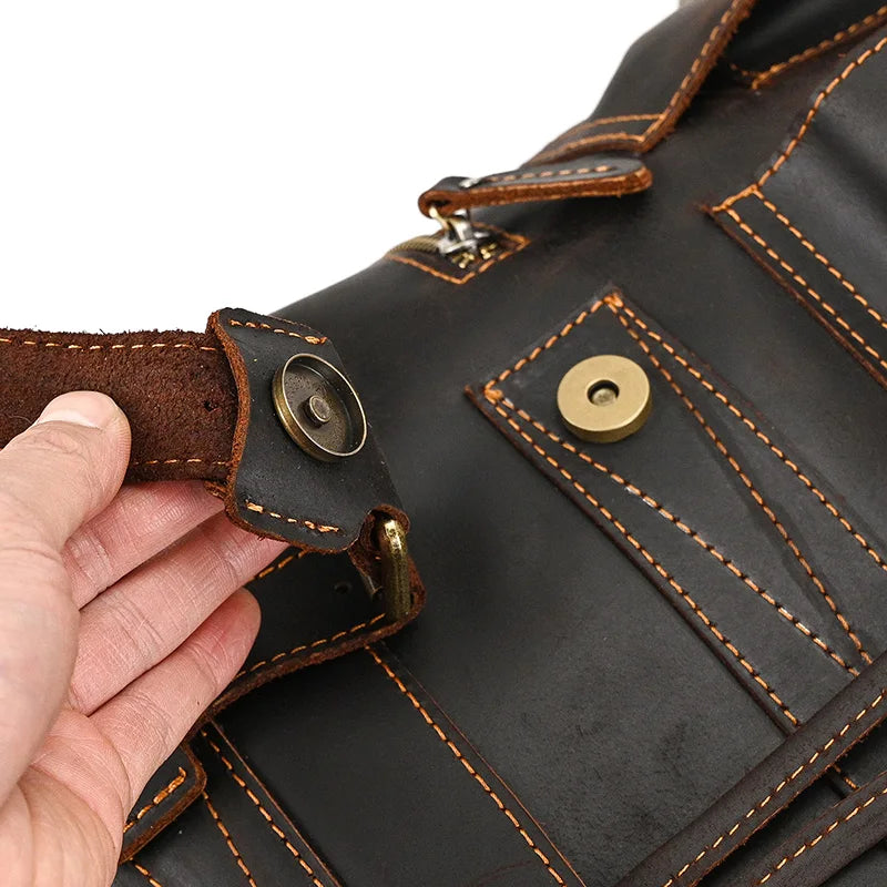 A close-up image of a person's hand holding the strap of a Genuine Leather Travel Backpack. The bag features orange stitching, brass hardware, and a front pocket with a snap button closure. The leather has a rugged, worn appearance with visible texture and character.