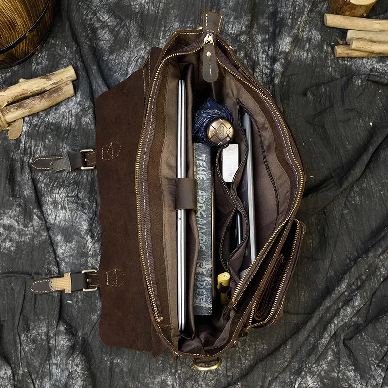 An overhead view of an open Leather 16" Business Briefcase reveals its contents: a laptop, a notebook, a watch, a smartphone, a pencil, and a rolled-up piece of clothing. The genuine cowhide leather bag is placed on a dark textured surface with pieces of wood in the background.