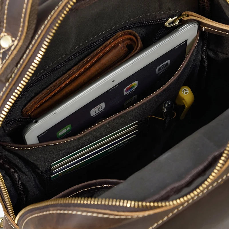 The Leather Travel Daypack Shoulder Bag, made from genuine cowhide leather, reveals its contents: a tablet, a brown wallet, several cards neatly placed in card slots, a pen, and a yellow key stored in a holder. This versatile daypack is also equipped with a dark fabric lining and a zipper closure.