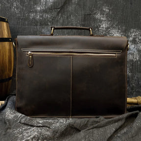 The Leather 16" Business Briefcase, crafted from genuine dark brown cowhide leather, is showcased against a textured dark backdrop. It boasts a top handle and an adjustable strap, with a large central compartment visible from the back and a pocket featuring a gold zipper. A partially visible wooden barrel appears to the left.