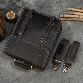 The Leather Business Travel Briefcase Backpack, a dark brown and versatile bag crafted from genuine cowhide leather with beige stitching and brass buckles, lies flat on a gray textured surface. It features adjustable straps and removable shoulder straps with snap hooks, while nearby wooden sticks and a log slice add a rustic touch.