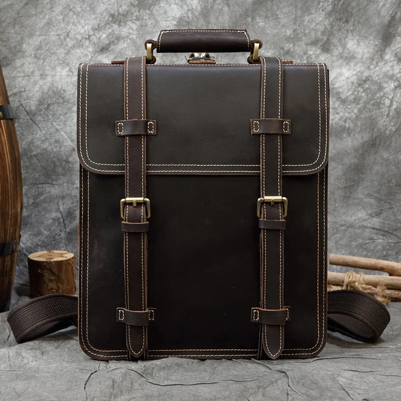 The Leather Business Travel Briefcase Backpack, made from genuine cowhide leather, stands upright against a textured gray background. It features a top handle and two front straps secured with brass buckles. The stitching details along the edges are prominently showcased as it rests on a concrete surface next to rustic wooden items.