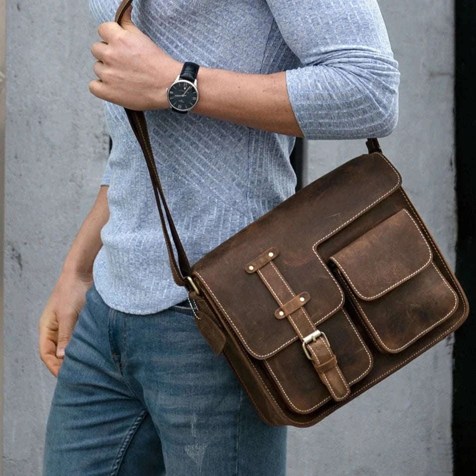 A person wearing a light gray, long-sleeve shirt and blue jeans is carrying a Leather Business Travel Shoulder Bag made from genuine cowhide leather, featuring multiple pockets and alloy hardware. They have a black wristwatch on their left wrist, standing against a plain, light-colored wall.