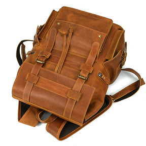 The Genuine Leather Travel Backpack, crafted from brown genuine leather, lies flat on a white surface. This vintage-inspired backpack features multiple compartments with buckled straps, a front pocket, padded shoulder straps for comfort, side pockets for additional storage, and a top handle for easy carrying.