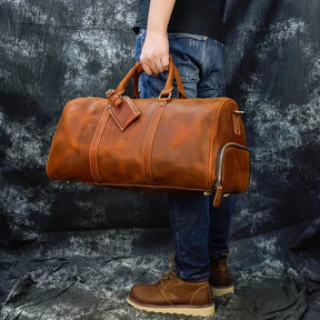 A person dressed in jeans and a black shirt is holding a large Weekender 24 Duffle Bag by its top handles. This leather bag features multiple compartments and buckles. The individual stands on a dark, textured surface with a mottled background, ideal for travelers.