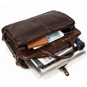 A Leather 16" Briefcase, crafted from genuine cow leather and partially open, reveals various items inside including a smartphone, a tablet, a leather wallet, a notebook, and what appears to be a laptop underneath. The brown briefcase features two handles and an adjustable strap buckle on the side.