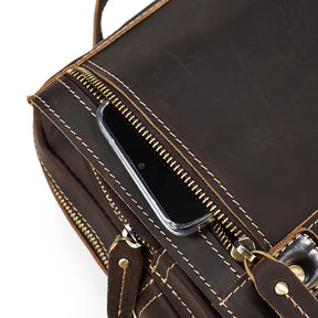 A close-up image of the Leather Travel Daypack Shoulder Bag made from dark brown genuine cowhide leather with beige stitching. The zipper is partially open, revealing a black smartphone inside. This versatile daypack features a metal hook and several compartments.