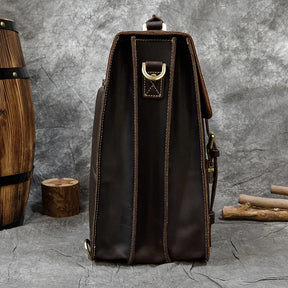 A side view of the Leather Business Travel Briefcase Backpack in dark brown genuine cowhide leather, showcasing stitched detailing and a brass ring strap attachment. The background features rustic elements, including a wooden barrel, a tree stump, and scattered wooden sticks, set against a textured gray backdrop.