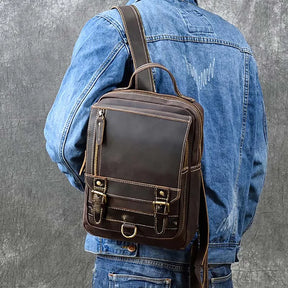 A person is wearing a blue denim jacket with the Leather Travel Daypack Shoulder Bag, made from genuine cowhide leather, slung over one shoulder. The bag features buckled straps, a front zipper pocket, and detailed stitching. The background is a textured gray.