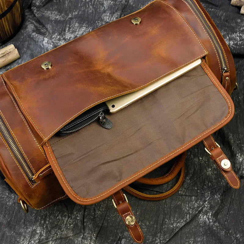 Introducing the Weekender 20 Leather Travel Bag: Crafted from genuine cowhide, this brown leather backpack features a front flap that opens to reveal an interior pocket with a partially visible tablet. The bag is equipped with brass hardware and side zipper pockets, all resting on a dark textured surface.