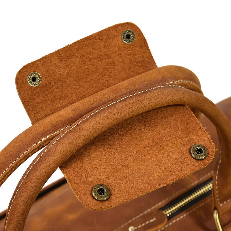 Close-up of a brown Weekender 24 Duffle Bag handle, showcasing detailed stitching and metal snap buttons on a small flap. Made from genuine cowhide leather, the texture is visible, and part of a zipper can be seen in the background—a perfect choice for travelers.