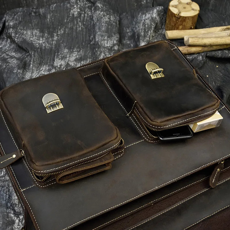 A close-up photo of the Leather 16" Business Briefcase showcasing its metal clasp closures. The briefcase, crafted from genuine cowhide leather, is dark brown with visible stitching and is displayed on a matching leather surface. Nearby, wooden logs and a textured black fabric background evoke an outdoor or rustic setting.