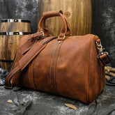 A large Weekender 18 Leather Bag crafted from genuine cowhide, featuring handles and an adjustable shoulder strap, rests on a dark fabric. Wooden barrels with black metal bands and a textured dark wall complete the rustic setting in the background, while its spacious interior pockets promise ample storage.