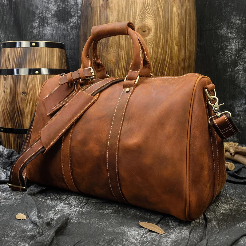 The Weekender 18 Leather Bag, a large brown duffel made of 100% genuine cowhide leather, is displayed on a textured surface. This stylish and functional bag boasts reinforced stitching, a padded shoulder strap, sturdy handles, and spacious interior pockets. Wooden barrels with metal bands in the background enhance the rustic setting.