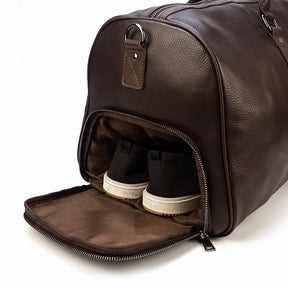 A dark brown Weekender Duffle 21 Leather Travel Bag with a side compartment unzipped, revealing a pair of black shoes with white soles neatly tucked inside. This sturdy duffle features silver hardware and a small ring attachment on the side.