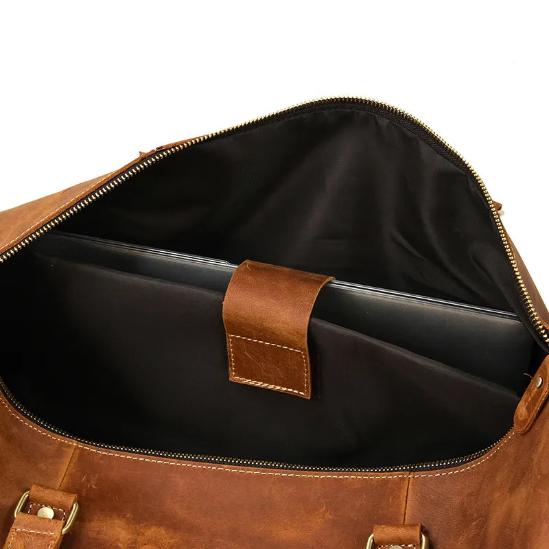 The Weekender 24 Duffle Bag, crafted from genuine cowhide leather, reveals a black laptop nestled in a padded compartment through its open zipper. The interior, lined with black fabric and featuring a brown leather strap for securing the laptop, makes it an ideal choice for travelers seeking a rustic, vintage look with metal hardware.