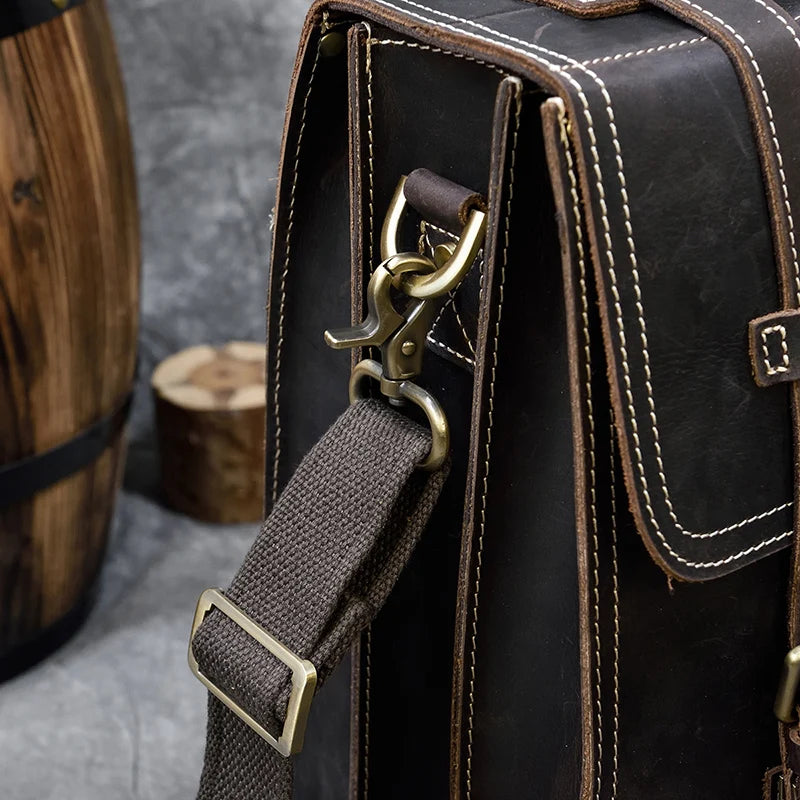 Close-up of the Leather Business Travel Briefcase Backpack in dark brown with white stitching, highlighting a sturdy metal clasp that fastens the woven fabric strap to the genuine cowhide leather bag. The backdrop includes a wooden barrel and block, creating a rustic setting ideal for business travel briefcases.