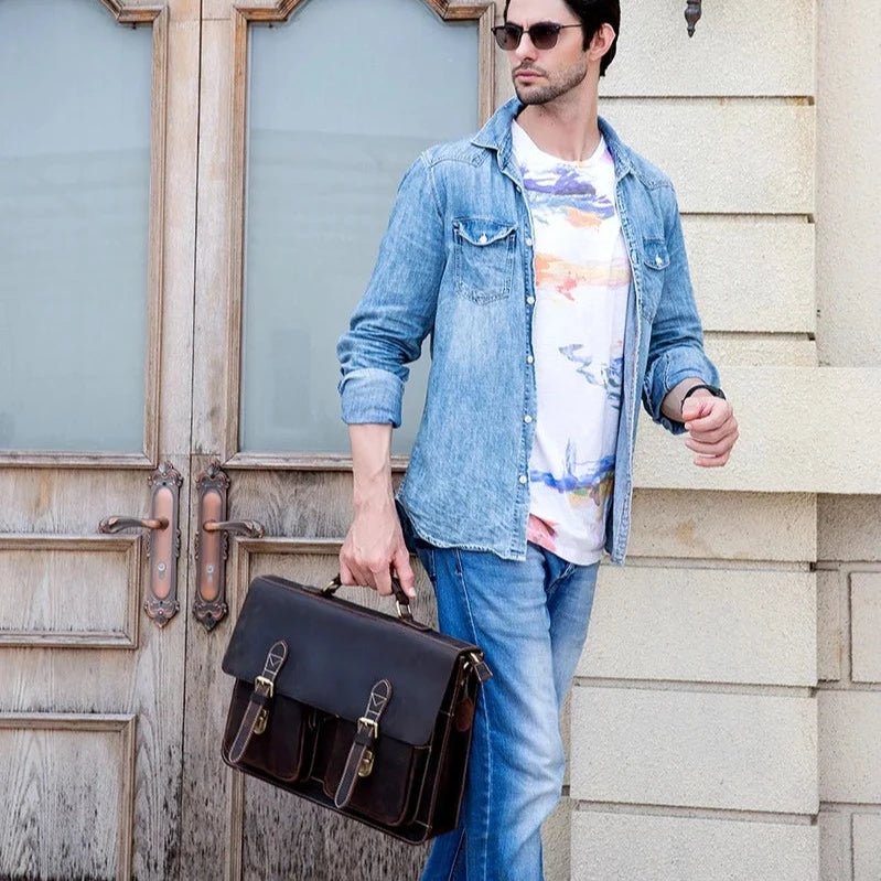 A man stands in front of a wooden door, wearing sunglasses, a light blue denim shirt over a white t-shirt with colorful patterns, and blue jeans. He holds the Leather 16" Business Briefcase made of genuine cowhide leather in his left hand and looks to his right.