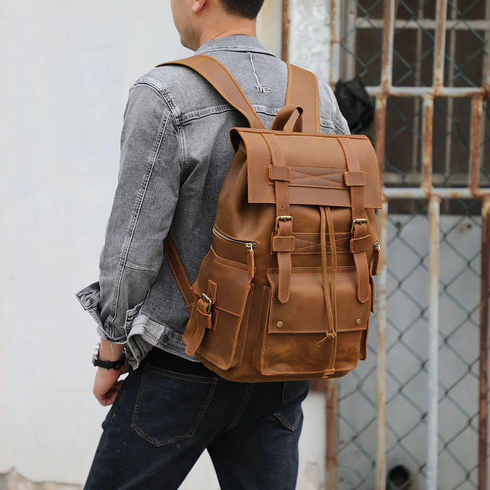 A man with short dark hair is sporting a grey denim jacket and dark jeans, carrying a Genuine Leather Travel Backpack in tan, which features multiple pockets and buckles. He stands in front of a weathered, metal-barred window with his back turned to the camera.