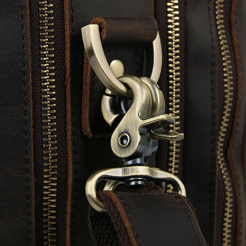 Close-up of a metallic clasp on the Leather 17" Business Travel Briefcase. The polished brass hardware is secured with a leather strap, and multiple gold-colored zippers run vertically along the surface next to the clasp. The bag also features an adjustable strap for comfort and convenience.