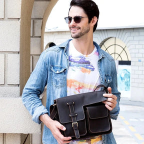 A man with dark hair sporting sunglasses, a blue denim jacket, and a vibrant graphic t-shirt grins as he holds his Leather Business Travel Shoulder Bag, meticulously crafted from genuine cowhide leather. He stands beside a building adorned with arched windows in the background.