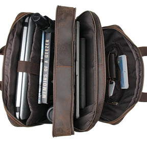 The Leather 17" Business Travel Briefcase is open, revealing a neatly organized interior. Inside, a laptop rests in its dedicated compartment alongside a book titled "Memoirs of a Geezer," pens, a water bottle, a smartphone, and various office supplies. The compartments are well-structured and come with an adjustable strap for convenience.