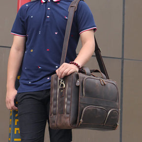 A person is wearing a navy blue polo shirt adorned with small, scattered red and yellow designs paired with black pants, while carrying a large Leather 17" Business Travel Briefcase featuring an adjustable strap. In the background, part of a tall building is visible.