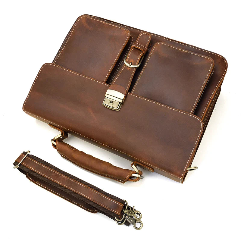 The Leather 16" Business Briefcase, crafted from brown genuine cowhide leather, is displayed with a stitched handle, front pockets, and a metal clasp. Beside it lies an adjustable shoulder strap featuring metal hooks and a padded section. The briefcase's interior and accessory attachment points are also visible.