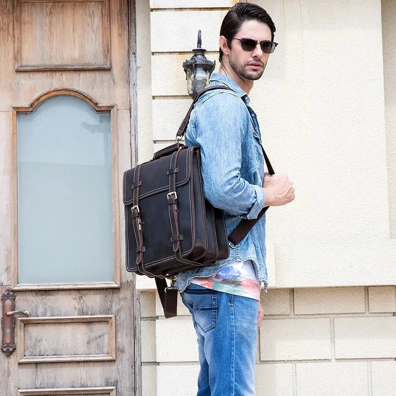 A man wearing sunglasses, a light blue denim shirt, loose-fitting jeans, and carrying the Leather Business Travel Briefcase Backpack in dark brown genuine cowhide leather on his back stands next to a wooden door and beige wall. He looks away from the camera.